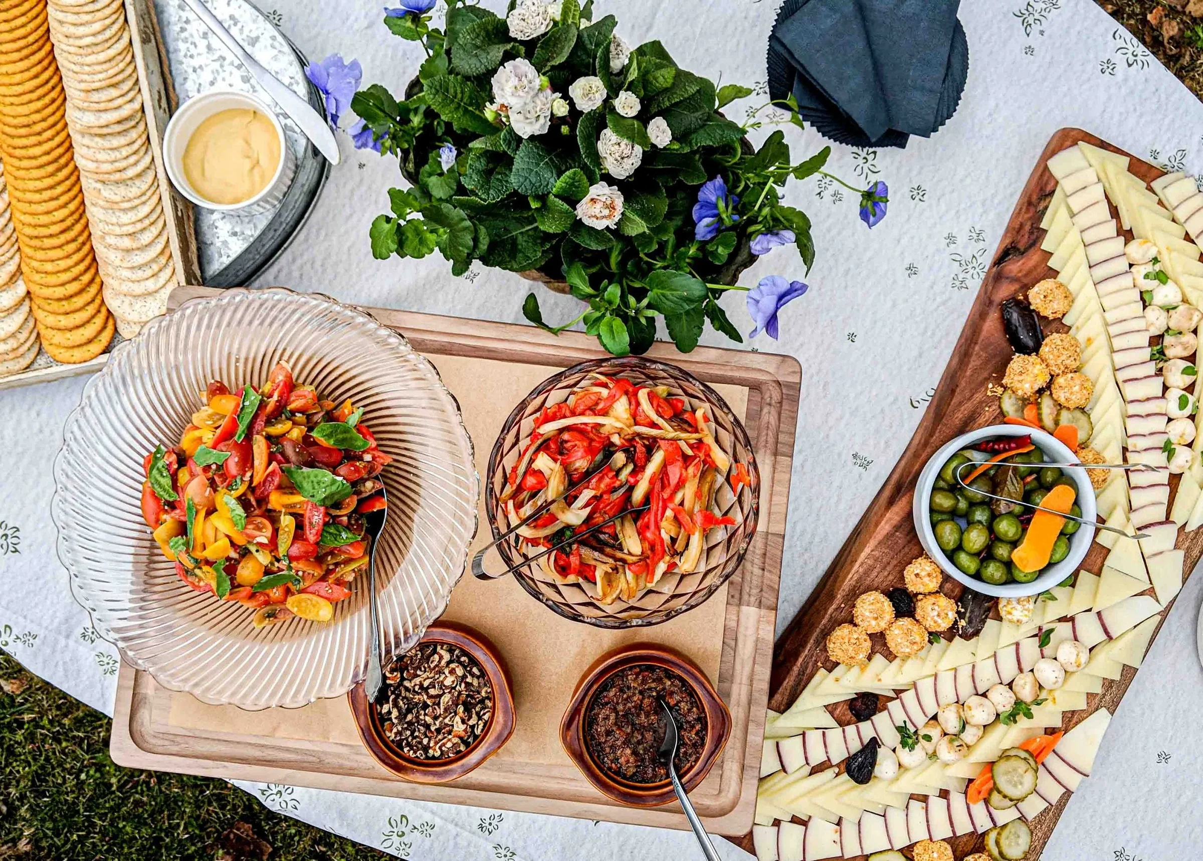 Overhead view of a catering table with a tomato and basil salad, roasted peppers and fennel, a cheese and olive board, crackers with hummus, and a small bouquet of flowers