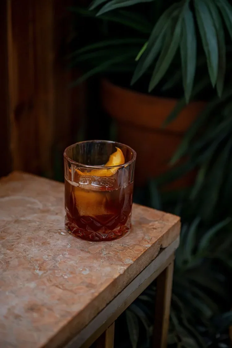 An old fashioned with a large ice cube and an orange peel in a cut-crystal rocks glass, set on a pink stone table in front of dark green plants