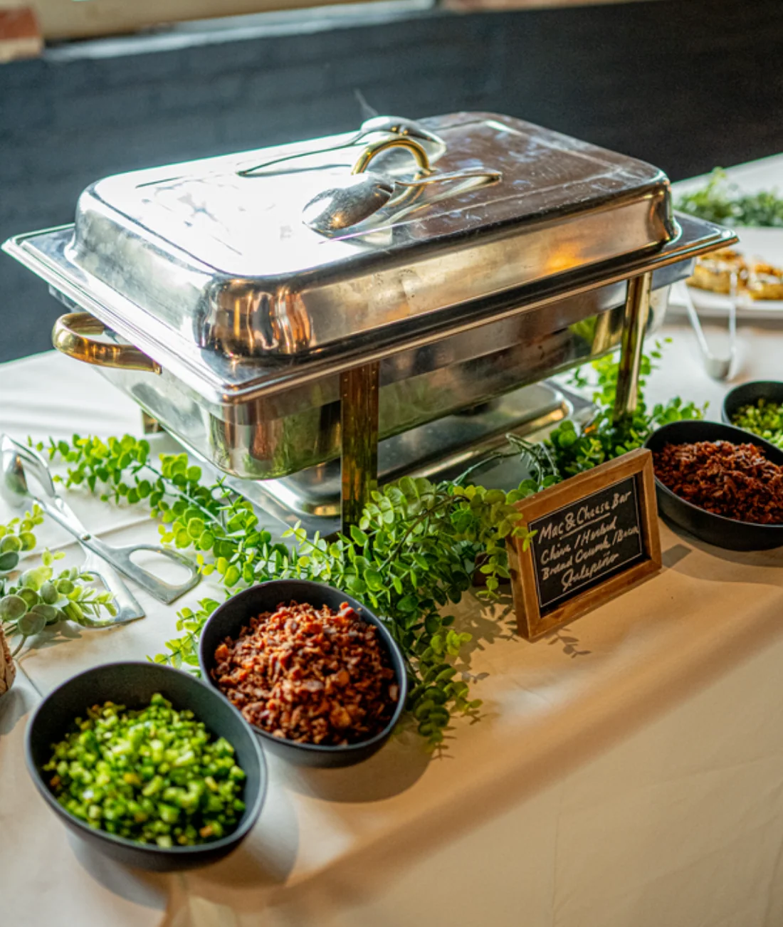 A mac and cheese bar on a linen-covered table, with a chafing dish, a small chalkboard sign, and black bowls of chives, bacon, and jalapeño
