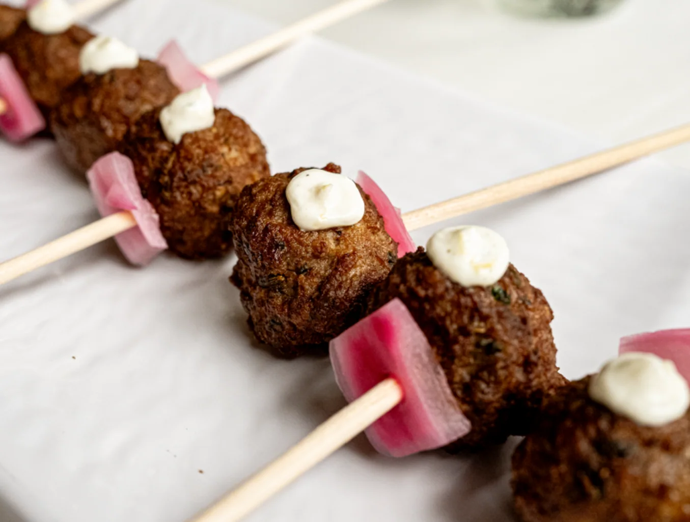 Skewers of golden meatballs with a dollop of cream and a piece of pickled pink onion each, arranged on white parchment