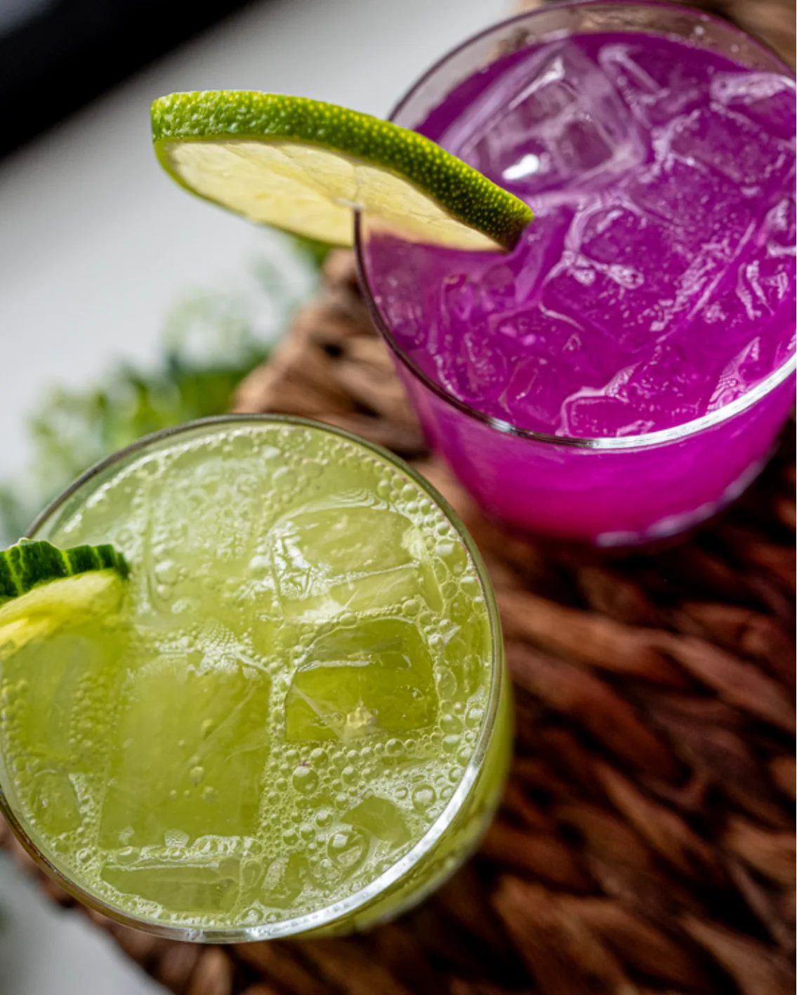Two iced zero proof cocktails seen from above, one bright magenta with a lime wheel and one pale green and sparkling with a cucumber slice, on a woven tray