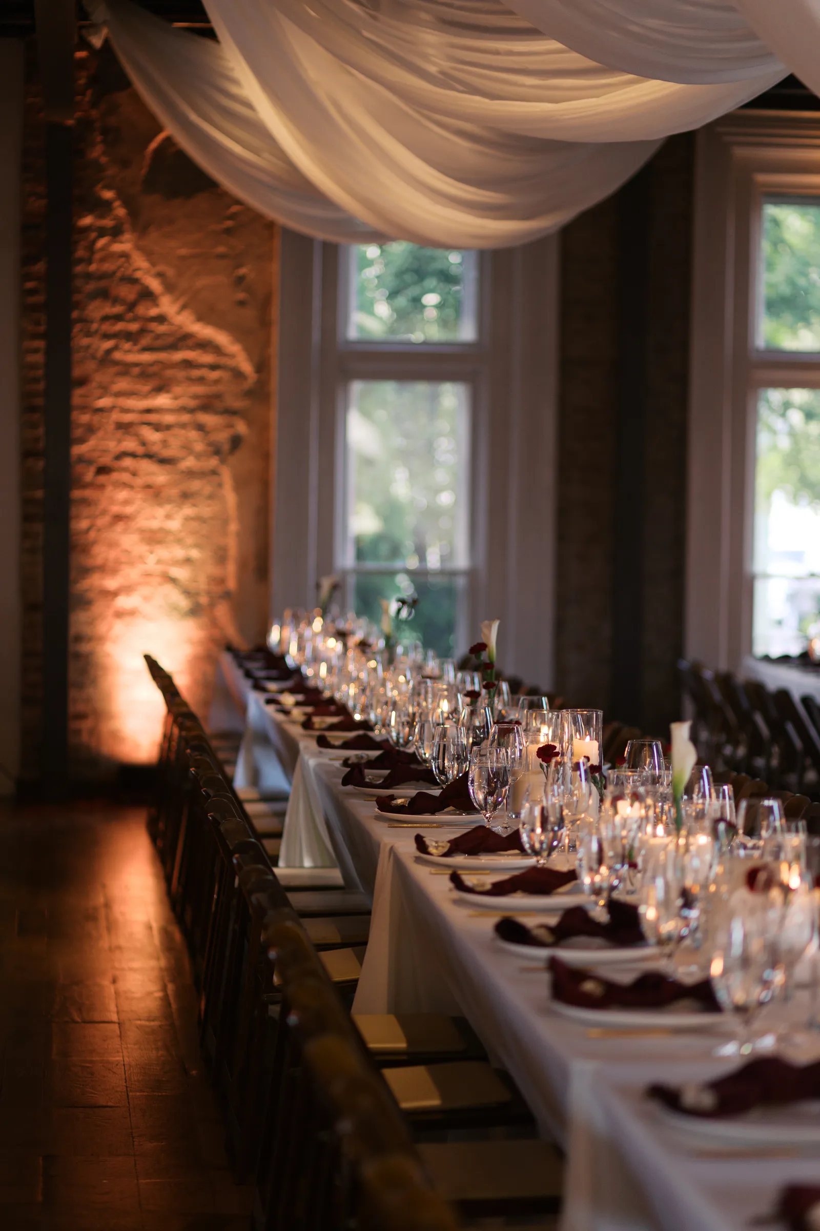 Long reception table lit with candles beneath draped fabric in a brick-walled hall