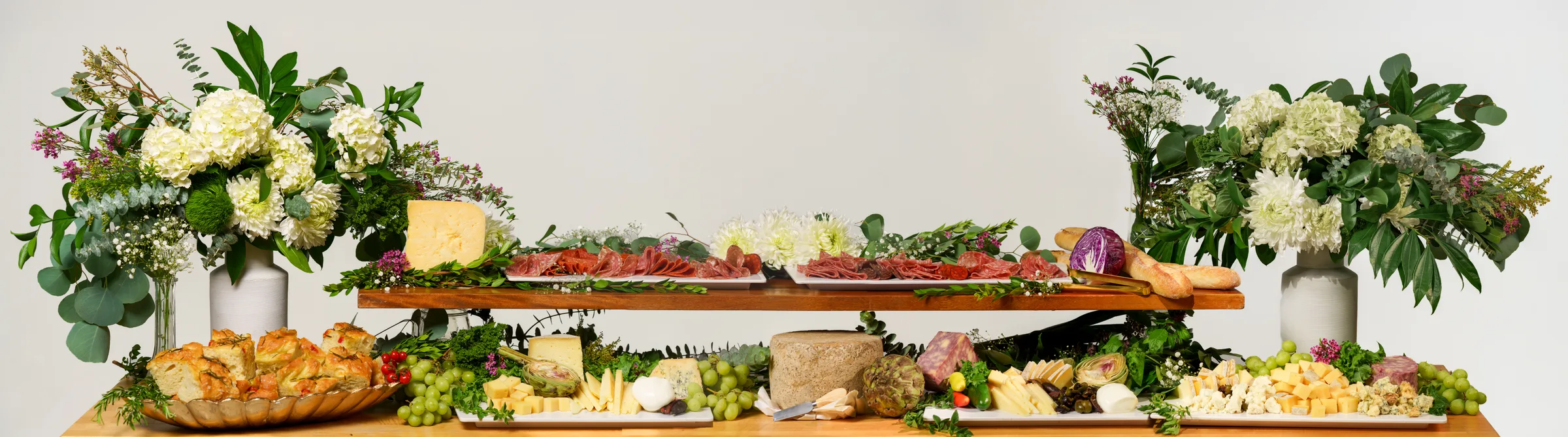 A grazing table of cured meats, cheeses, grapes, and bread between tall floral arrangements